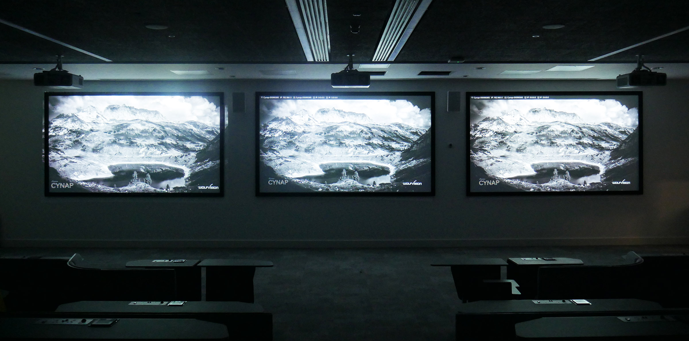 three large projection screens at the front of a darkened university lecture theatre