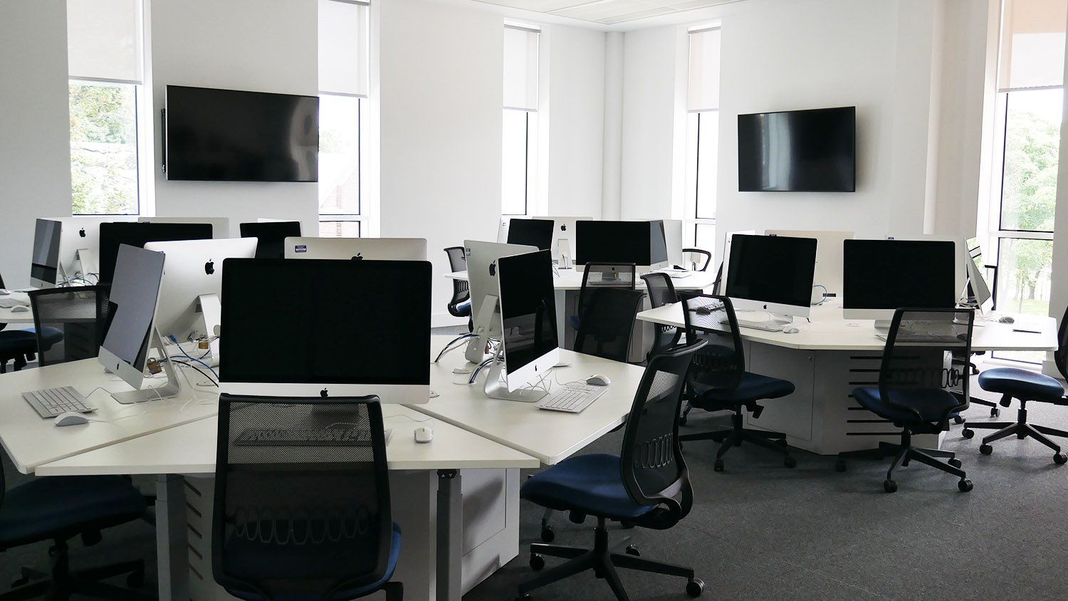 classroom with clusters of desks with iMac computers and screens on the walls